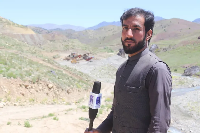 An Afghan journalist holding a microphone reports from a mountain valley
